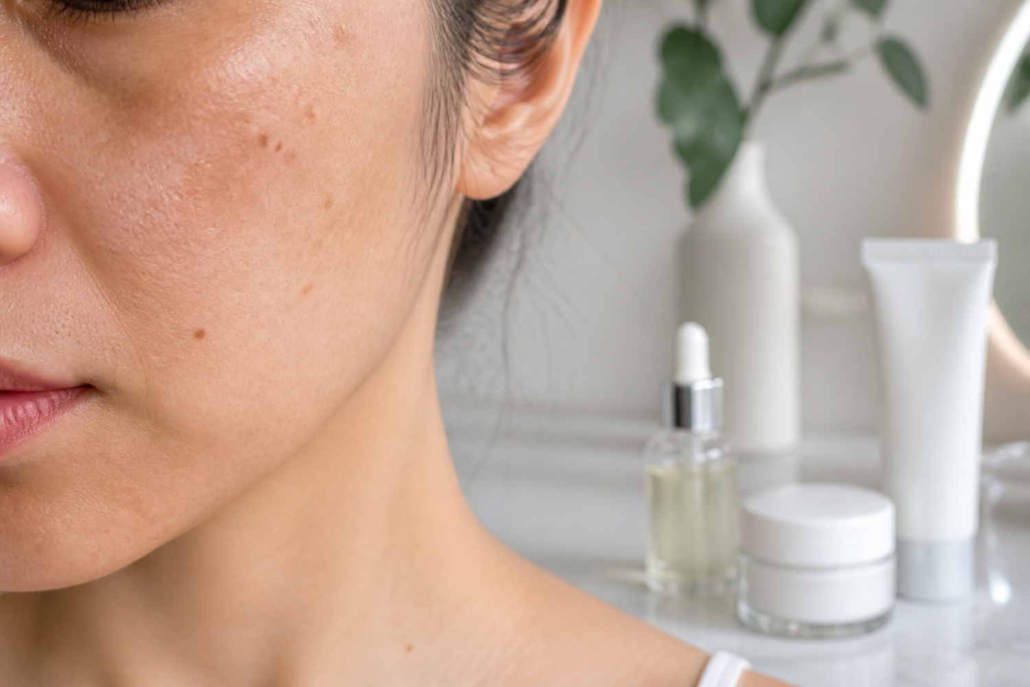 Close-up of facial skin with dark spots next to serum, moisturizer, and sunscreen on a vanity